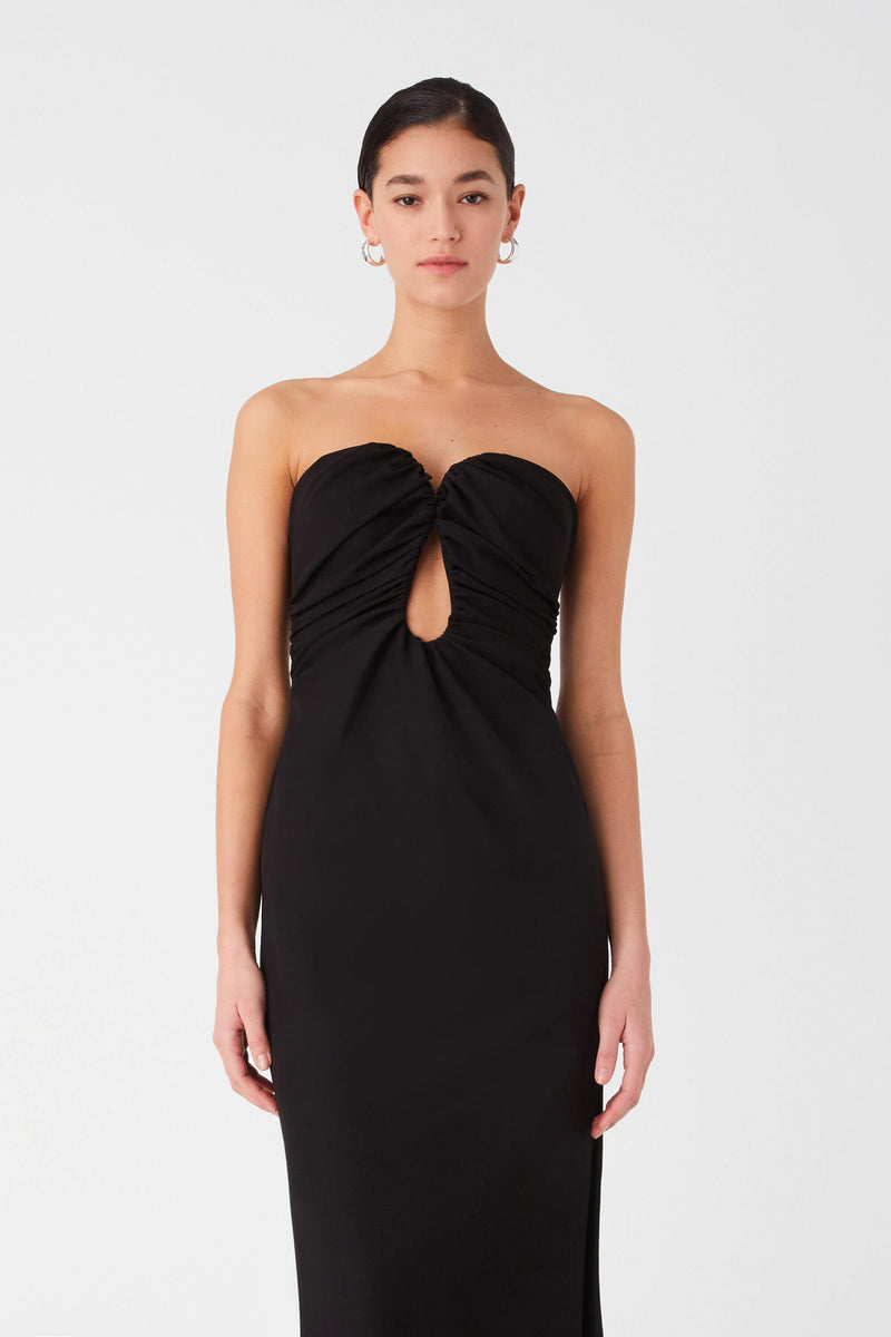 Woman posing in a black strapless gown with a front cut-out detail. Dress is maxi length with ruching around bust and curved strapless neckline.