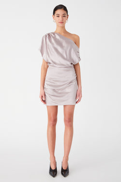 woman models a stylish of the shoulder satin mini dress, worn with earrings and black pointed heels. Dress is silver with oversized shoulder draping, and body-con pencil skirt.