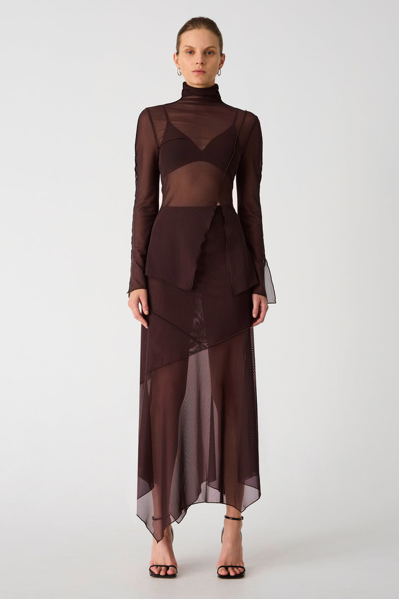 Girl wearing brown mesh top and maxi skirt set. Top has high neck and long sleeves, with an asymmetrical hemline and cutout. Skirt is asymmetrical, both pieces feature exposed seem detailing and are semi sheer.