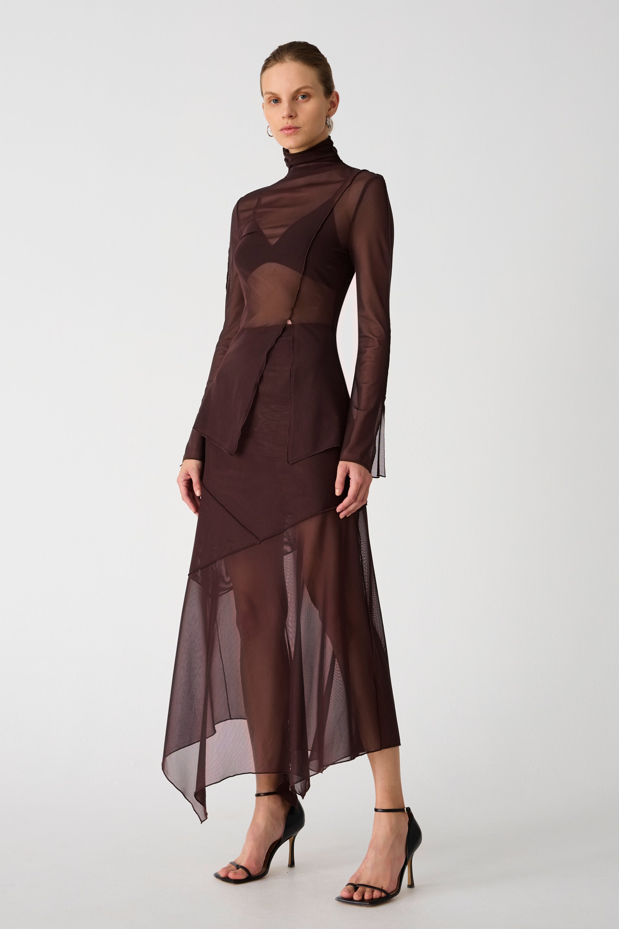 Girl wearing brown mesh top and maxi skirt set. Top has high neck and long sleeves, with an asymmetrical hemline and cutout. Skirt is asymmetrical, both pieces feature exposed seem detailing and are semi sheer.