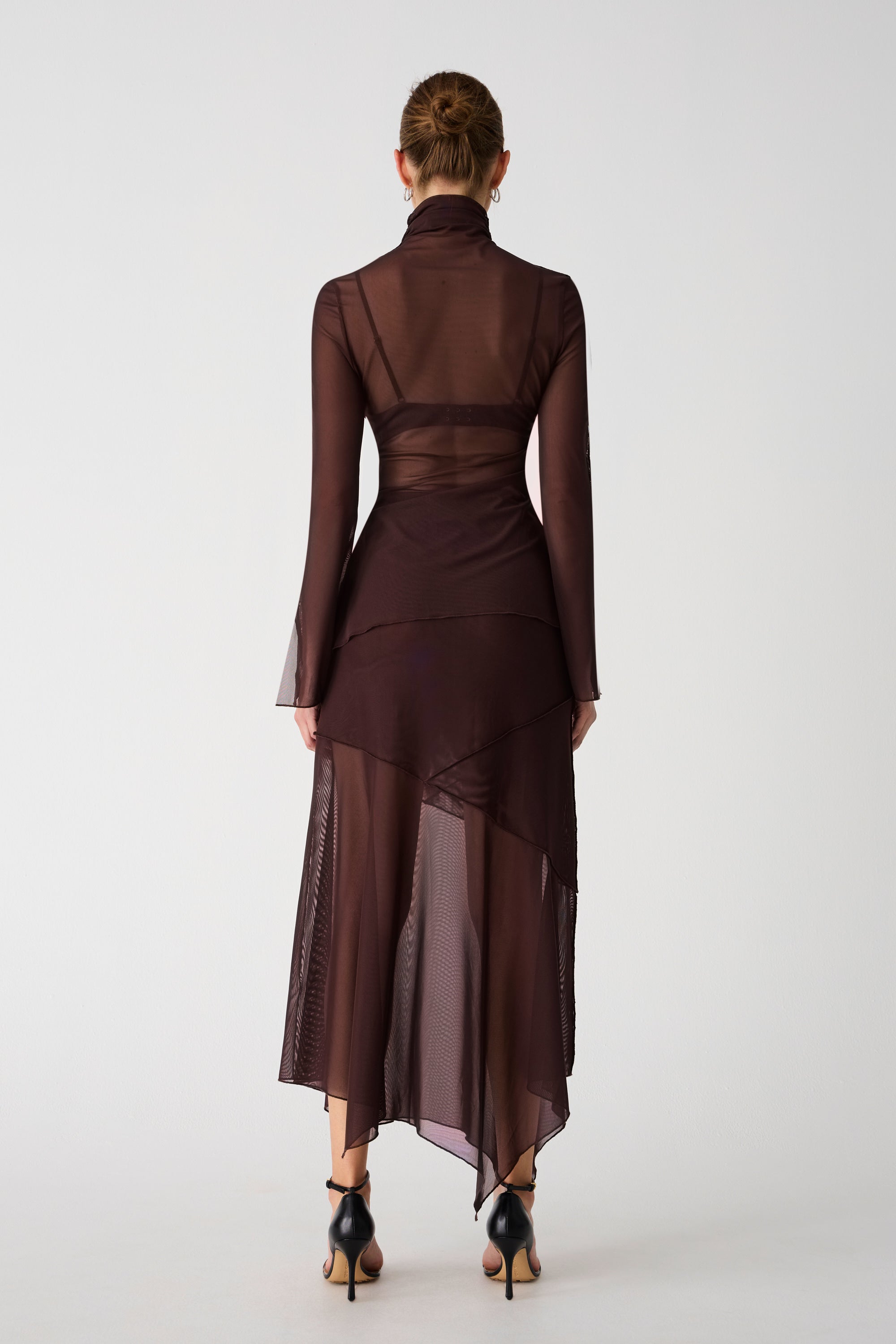 Girl wearing brown mesh top and maxi skirt set. Top has high neck and long sleeves, with an asymmetrical hemline and cutout. Skirt is asymmetrical, both pieces feature exposed seem detailing and are semi sheer.