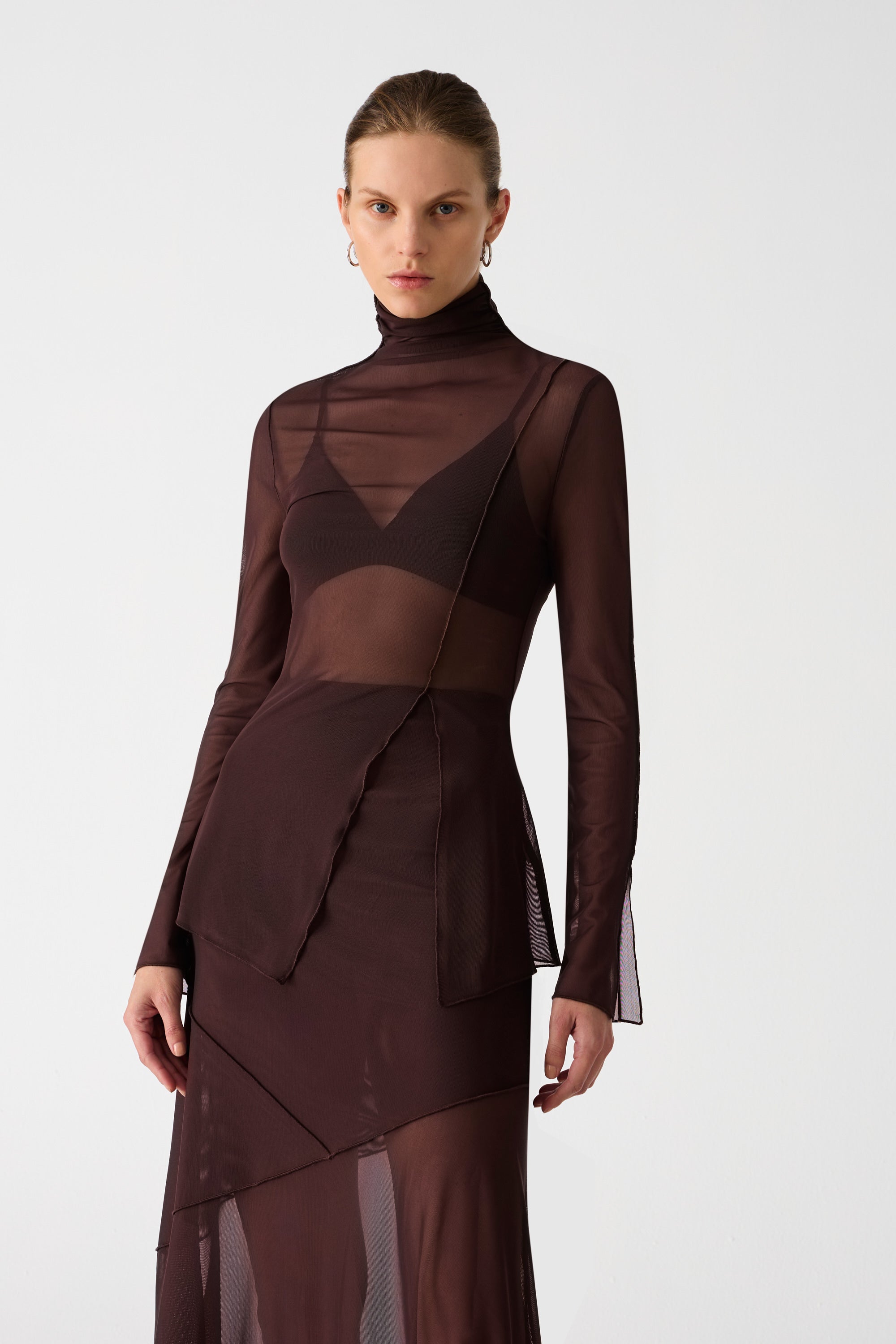 Girl wearing brown mesh top and maxi skirt set. Top has high neck and long sleeves, with an asymmetrical hemline and cutout. Skirt is asymmetrical, both pieces feature exposed seem detailing and are semi sheer.