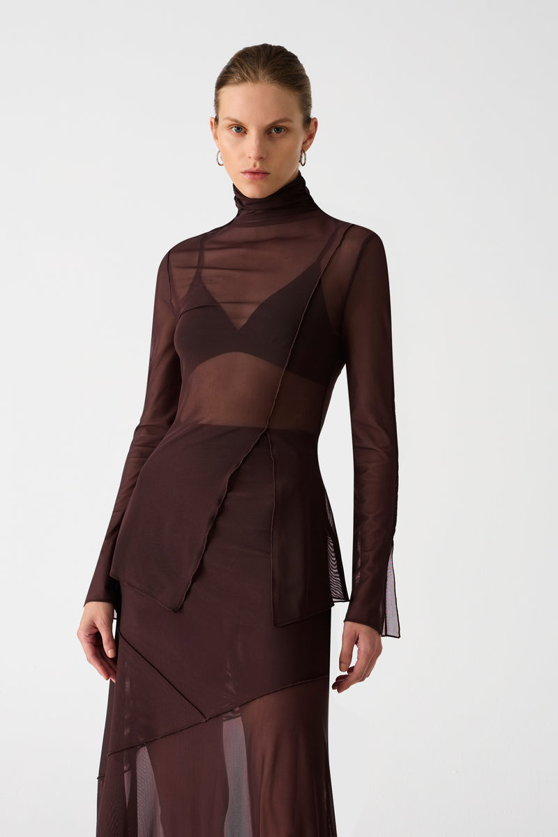 Girl wearing brown mesh top and maxi skirt set. Top has high neck and long sleeves, with an asymmetrical hemline and cutout. Skirt is asymmetrical, both pieces feature exposed seem detailing and are semi sheer.