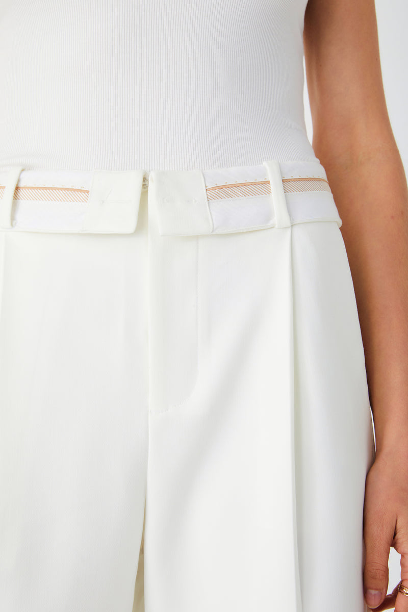 Close up of white pant waistband. Detailing fold over waist band and front pleats. Worn with white tank top.