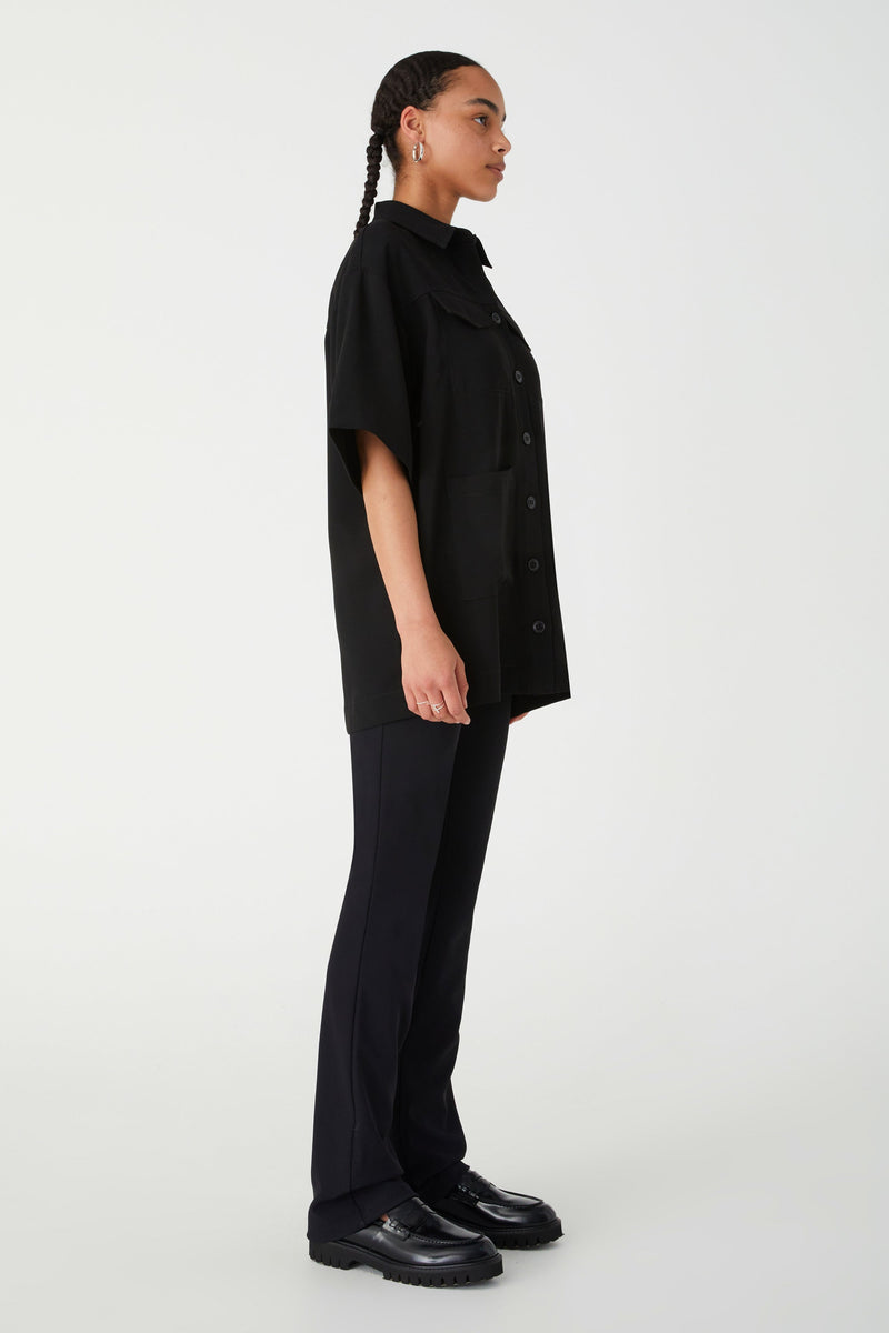Girl wearing black oversized button down shirt with black flared pants and black shoes. Shirt has oversized pockets and short sleeves.