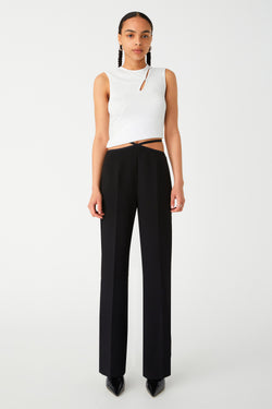 A woman in a chic white sleeveless top and black high-waisted trousers posing against a plain background. Pants have asymmetrical waist band, with two waist straps on either side, creating a diamond shape in the centre.
