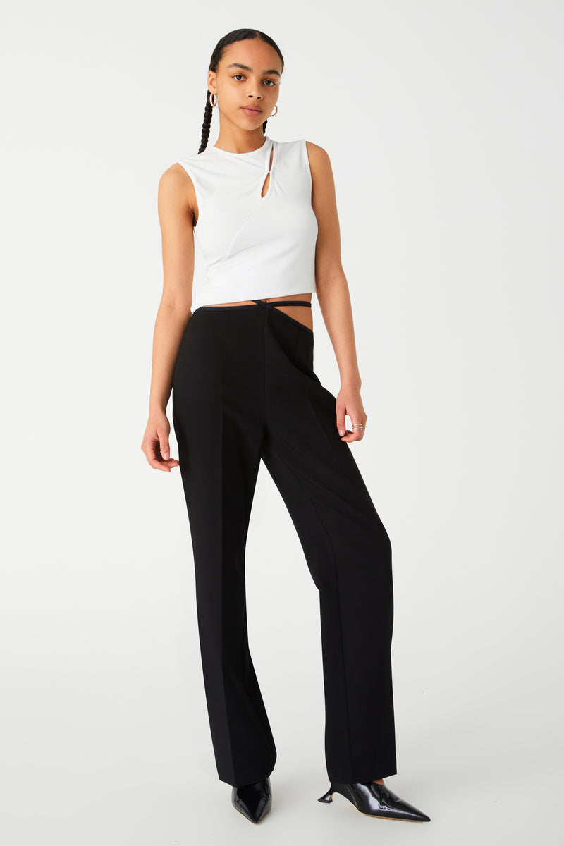 A woman in a chic white sleeveless top and black high-waisted trousers posing against a plain background. Pants have asymmetrical waist band, with two waist straps on either side, creating a diamond shape in the centre.