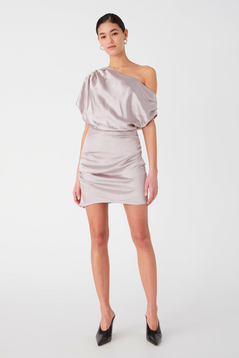woman models a stylish of the shoulder satin mini dress, worn with earrings and black pointed heels. Dress is silver with oversized shoulder draping, and body-con pencil skirt.