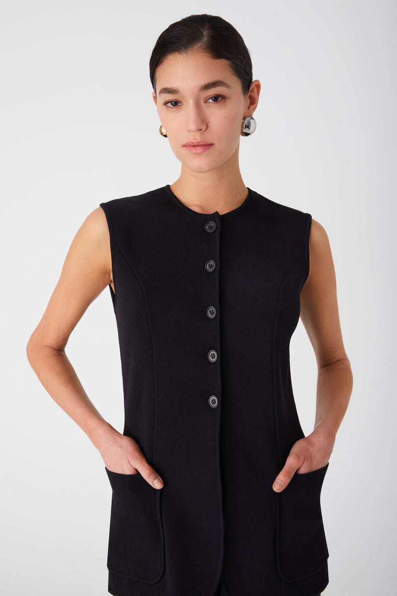 Girl wearing high neck knit vest top with black pants and black heels. Vest top is long line, with buttons going half way down, ending below the bust. Vest top is a knit material and has two large pockets on either side near the hips.