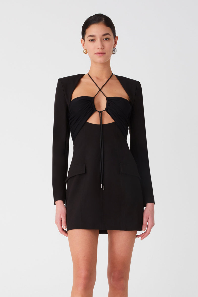 Woman in black mini dress with cut-out details and high heels. Dress has long sleeves and pockets, with a tie up detail around the neck connecting the bust cups.