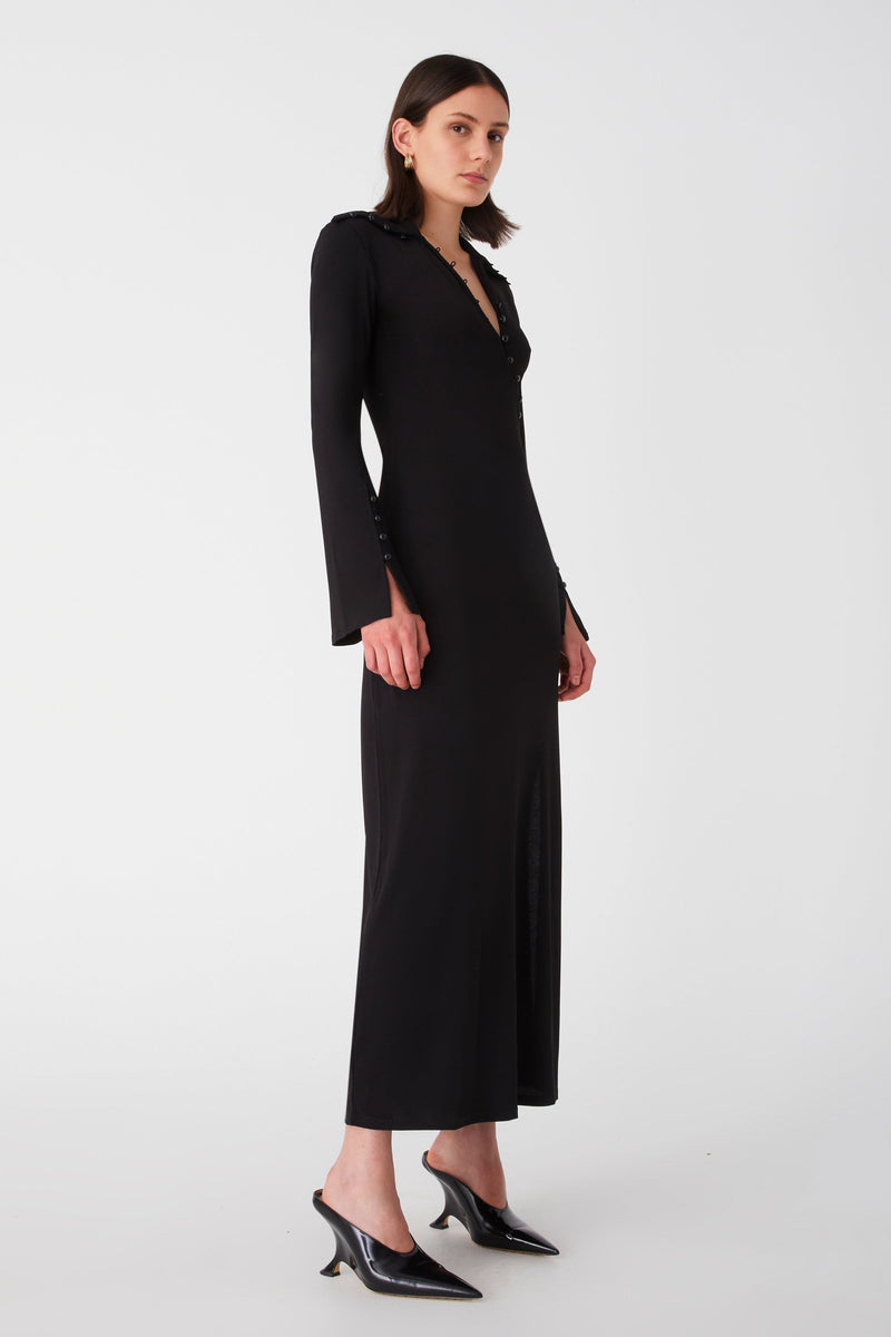 Woman in ankle length black dress with a unique diagonal button detail and long sleeves, paired with classic black heels. Dress has button detailing on sleeves, and an exaggerated collar.