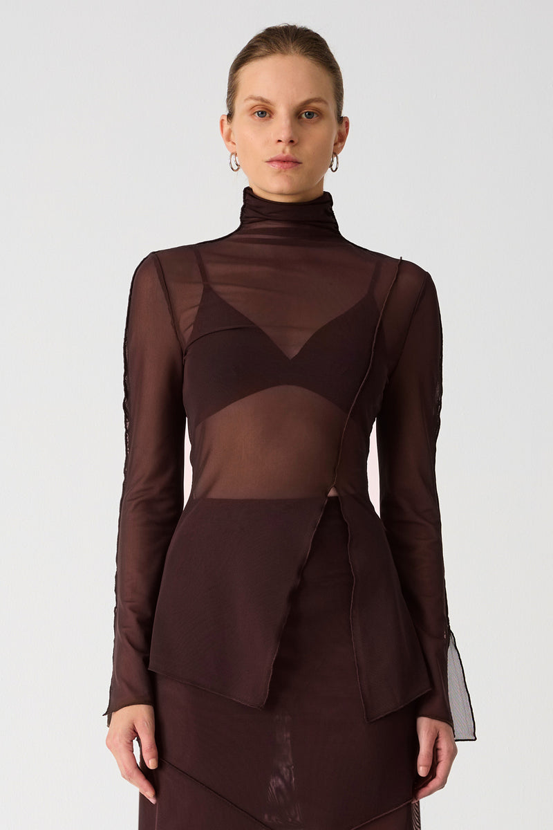 Girl wearing brown mesh top and maxi skirt set. Top has high neck and long sleeves, with an asymmetrical hemline and cutout. Skirt is asymmetrical, both pieces feature exposed seem detailing and are semi sheer.